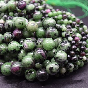 May include: A close-up of a pile of green, black, and pink gemstone beads. The beads are round and have a smooth, polished finish.