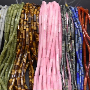 May include: Multiple strands of cylindrical gemstone beads in various colours including olive green, brown tiger eye, light pink, dark grey, royal blue, and reddish-orange. Each strand consists of smooth, uniform tube-shaped beads arranged vertically.