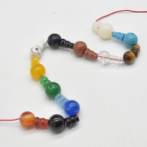 Op de afbeelding: Een kleurrijke kralenketting met een verscheidenheid aan edelsteen kralen in een regenboog van kleuren. De kralen omvatten oranje, zwart, blauw, groen, geel, wit en bruin. De ketting is geregen op een rood koord.