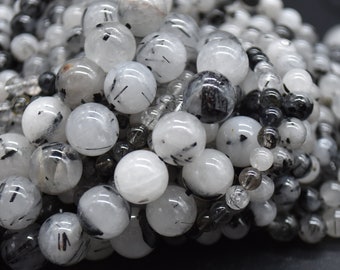 May include: Close-up of a collection of round beads in varying sizes. The beads are primarily white and clear, with black inclusions and markings. The beads are strung together, creating a textured, layered effect.