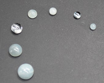 May include: A collection of round, smooth, light blue and clear cabochons of varying sizes arranged in a semi-circle on a dark gray surface.