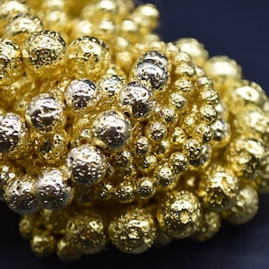 May include: A close-up of a pile of gold-coloured lava rock beads. The beads are round and have a rough, textured surface.