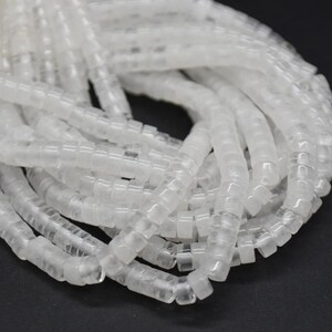 May include: Close-up of a pile of clear, square beads. The beads are strung together, forming multiple strands. The beads are translucent and appear to be made of plastic or glass. The background is a dark, solid colour.