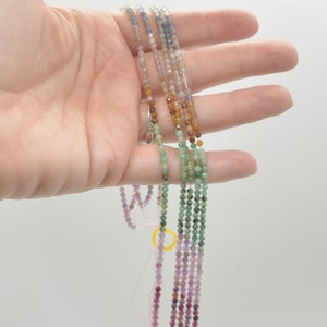 May include: A strand of gemstone beads in various colors, including purple, green, brown, and blue. The beads are small and round, and they are strung on a clear thread.