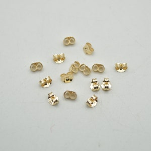 May include: A collection of small, shiny gold-colored earring backs scattered on a light-colored surface. The earring backs have a butterfly shape, with two loops for the earring post to slide through. The backs are made of a reflective metal.