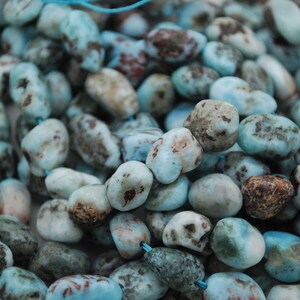 May include: Close-up of a collection of tumbled, irregular-shaped beads. The beads are mainly light blue with brown and white markings. They are strung together on a thin, light blue cord, suggesting they are for jewellery making.