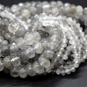 Könnte beinhalten: Eine Strähne aus durchscheinenden, facettierten, grauen Quarzperlen. Die Perlen sind rund und haben eine leicht unregelmäßige Form. Die Perlen sind auf einem durchsichtigen Faden aufgefädelt.
