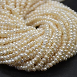 Op de afbeelding: Een streng kleine, crèmekleurige zoetwaterparels. De parels zijn rond en hebben een licht onregelmatige vorm. Ze zijn aan elkaar geregen op een doorzichtige draad.