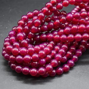 May include: A collection of smooth, round, deep magenta-coloured beads strung together in multiple strands. The beads have a glossy, semi-translucent finish, reflecting light across their spherical surfaces, creating a dense, uniform cluster of vibrant red-toned jewellery components.