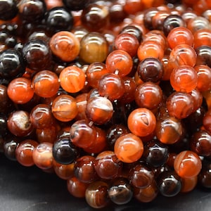 Pode incluir: Um close-up de um fio de contas de cornalina. As contas são uma mistura de cores vermelho, laranja e marrom com um padrão de ágata natural. As contas são redondas e lisas.