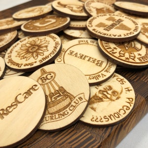 May include: A collection of round wooden tokens with various designs and text, including "ResCare", "BC Curling Club", "Sauk Valley", "Believe", "Play It Safe", "Caring for Each Other and Our Patients", and "Radiology".