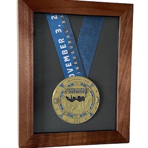 May include: A framed medal from the Las Vegas Marathon, featuring a gold-colored medal with a blue ribbon. The medal is displayed in a wooden frame with a black background. The ribbon has the text "November 3, 2 Inaugural Ra". The medal has the text "Las Vegas Marathon" and "26.2".