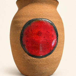May include: A small, rounded ceramic vase with a textured, matte tan exterior. The vase features a prominent, circular, glossy red accent patch on the front, which is outlined by a dark, charcoal-colored border. The rim is also dark and smooth.