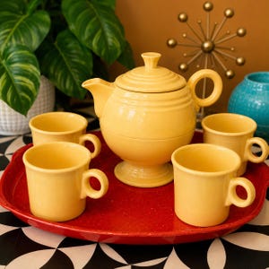 May include: A bright yellow ceramic teapot with a matching lid and four yellow mugs arranged on a large, round, speckled red serving tray. The teapot features a pedestal base, a curved handle, and a spout, while each mug has a circular handle.