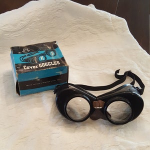 May include: Vintage black Cover Goggles with clear lenses and a brown nose guard. The goggles are on a white fabric surface with a blue and white box that says "Cover Goggles" and "Work safely - Protect your eyes".