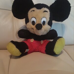 May include: A plush Mickey Mouse toy with black ears, a black nose, and large white eyes. The toy is wearing red shorts with white polka dots and has yellow feet.