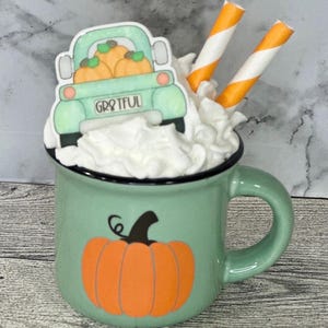 May include: A green mug with a black rim, filled with whipped cream and topped with a truck carrying pumpkins. The mug has a pumpkin graphic and the word "Grateful" on the truck. Two orange and white straws are in the whipped cream.