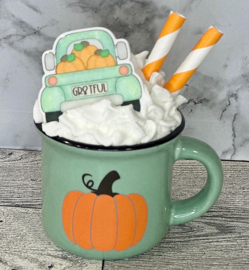 May include: A green mug with a black rim, filled with whipped cream and topped with a truck carrying pumpkins. The mug has a pumpkin graphic and the word "Grateful" on the truck. Two orange and white straws are in the whipped cream.
