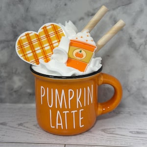 May include: An orange mug with the words "PUMPKIN LATTE" in white. The mug is filled with whipped cream, a heart-shaped decoration with an orange plaid pattern, and a pumpkin spice latte decoration. Two wafer sticks are placed on top.