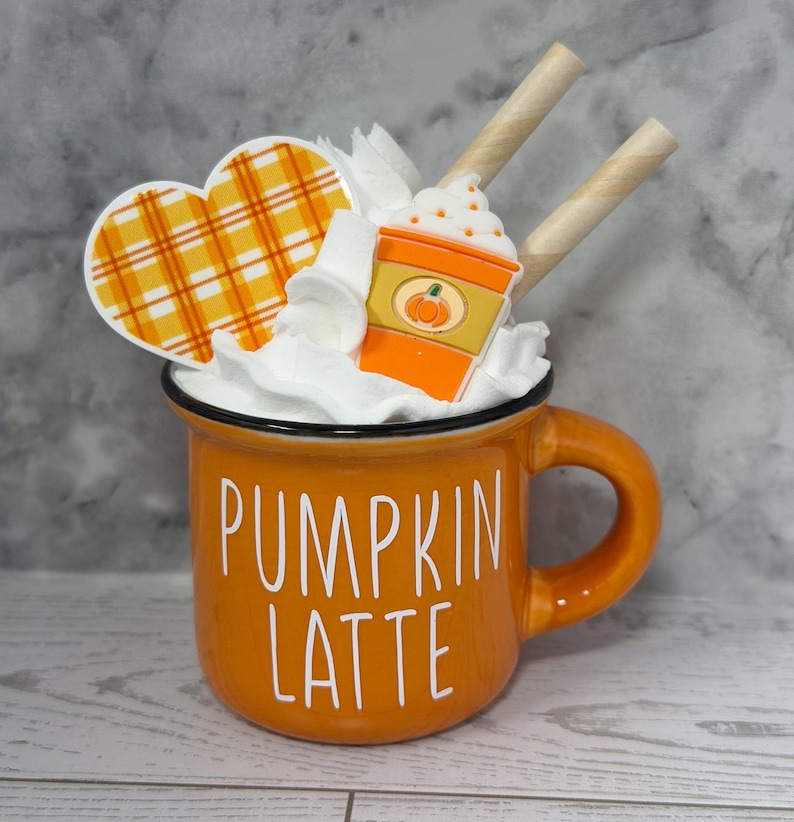 May include: An orange mug with the words "PUMPKIN LATTE" in white. The mug is filled with whipped cream, a heart-shaped decoration with an orange plaid pattern, and a pumpkin spice latte decoration. Two wafer sticks are placed on top.