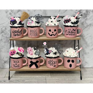 May include: Eight pink ceramic mugs decorated with Halloween themes. The mugs are arranged on a two-tiered black metal stand. The mugs feature various Halloween designs, including witches, pumpkins, ghosts, and spiders. The text on the mugs includes "Witches Brew", "Magic Potion", "It's October Witches", "Trick or Treat", and "Spooky".