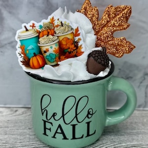 May include: A white ceramic mug with a black handle and the text "But First, Coffee" in a black, bold font. The mug is on a white surface.