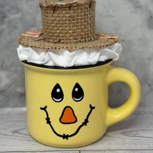 May include: A yellow ceramic mug with a painted scarecrow face, featuring black eyes, an orange nose, and a smiling mouth. The mug is topped with a small, woven straw hat with a white ruffled trim. The handle is yellow.