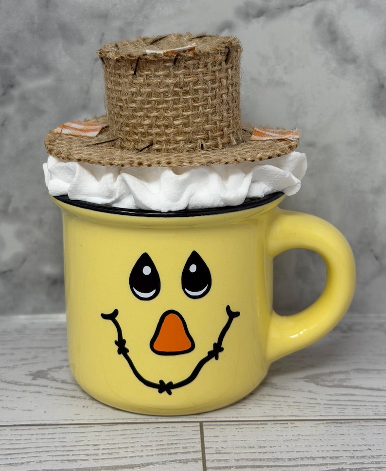 May include: A yellow ceramic mug with a painted scarecrow face, featuring black eyes, an orange nose, and a smiling mouth. The mug is topped with a small, woven straw hat with a white ruffled trim. The handle is yellow.