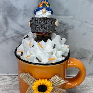 May include: An orange ceramic mug decorated with a sunflower and a burlap bow. The mug is filled with white whipped cream and topped with sprinkles. A gnome figure holding a sign that says "Happy Fall" sits on top.