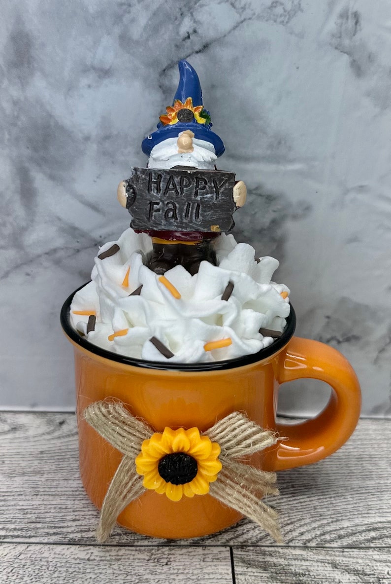 May include: An orange ceramic mug decorated with a sunflower and a burlap bow. The mug is filled with white whipped cream and topped with sprinkles. A gnome figure holding a sign that says "Happy Fall" sits on top.