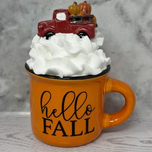 May include: Orange ceramic mug with a black rim and handle, featuring the text "hello FALL" in black script. The mug is topped with white decorative fluff and a miniature red truck carrying pumpkins.