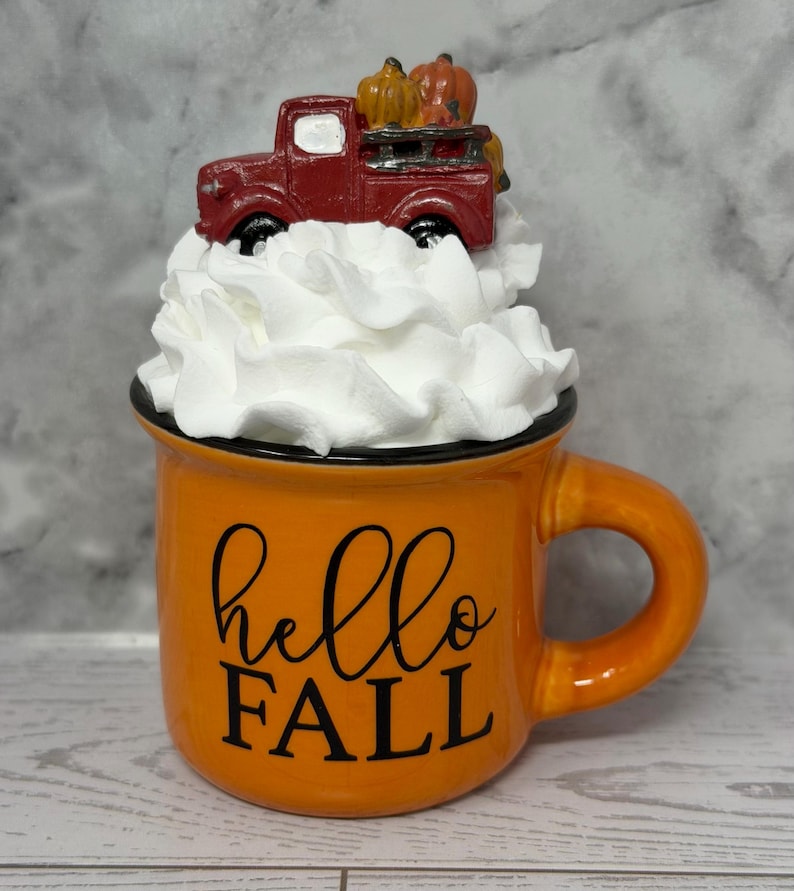 May include: Orange ceramic mug with a black rim and handle, featuring the text "hello FALL" in black script. The mug is topped with white decorative fluff and a miniature red truck carrying pumpkins.