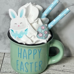 Op de afbeelding: Een groene keramische mok met een witte handgreep en de tekst "Happy Easter" in blauwe letters. De mok is gevuld met slagroom, een blauwe en witte konijnenvormige koek en een blauwe en roze eivormige koek. Twee blauw-wit gestreepte rietjes steken uit de slagroom.