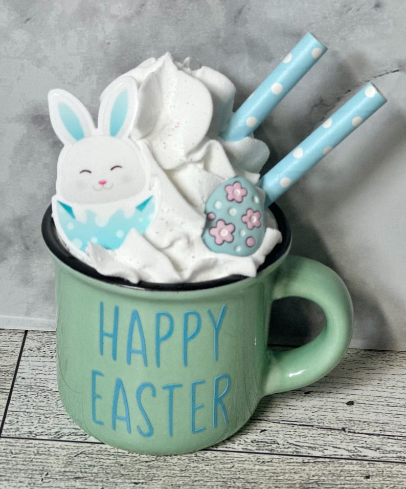Op de afbeelding: Een groene keramische mok met een witte handgreep en de tekst "Happy Easter" in blauwe letters. De mok is gevuld met slagroom, een blauwe en witte konijnenvormige koek en een blauwe en roze eivormige koek. Twee blauw-wit gestreepte rietjes steken uit de slagroom.
