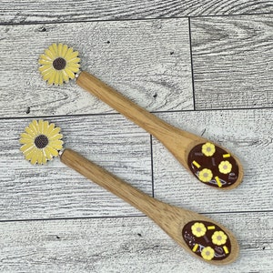 May include: Two wooden spoons with yellow sunflower designs on the handles. Each spoon is filled with dark brown chocolate and yellow sprinkles.