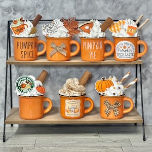 May include: A two-tiered shelf displays six orange mugs with white whipped cream, cinnamon sticks, and decorative accents. Each mug has a different label, including "Pumpkin Spice" and "Pumpkin Pie". The mugs are decorated with bows and fall-themed toppers.
