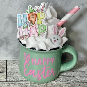 Op de afbeelding: Een groene keramische mok met witte slagroom, een roze-wit gestreepte rietje, een konijnengezicht en een "HOP HOP" bordje versierd met wortels en bloemen. De mok heeft de tekst "HAPPY Easter" in roze.