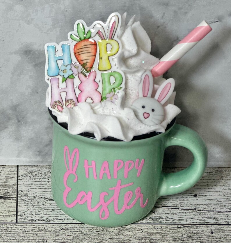 Op de afbeelding: Een groene keramische mok met witte slagroom, een roze-wit gestreepte rietje, een konijnengezicht en een "HOP HOP" bordje versierd met wortels en bloemen. De mok heeft de tekst "HAPPY Easter" in roze.
