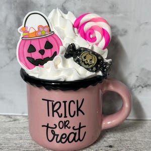 May include: Pink ceramic mug with a black rim and handle. The mug has white whipped cream on top with a pink candy cane, a pink pumpkin with a candy corn bucket, and a black and gold candy with a pumpkin face. The mug says "Trick or Treat" in black lettering.