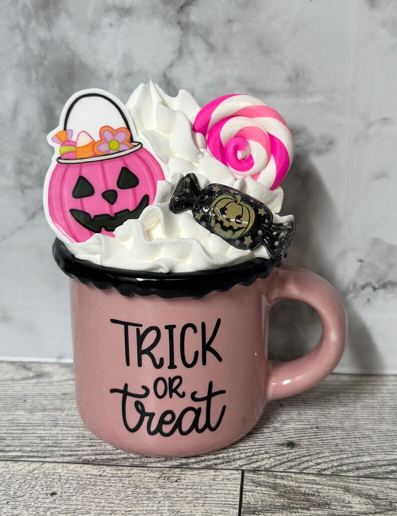 May include: Pink ceramic mug with a black rim and handle. The mug has white whipped cream on top with a pink candy cane, a pink pumpkin with a candy corn bucket, and a black and gold candy with a pumpkin face. The mug says "Trick or Treat" in black lettering.