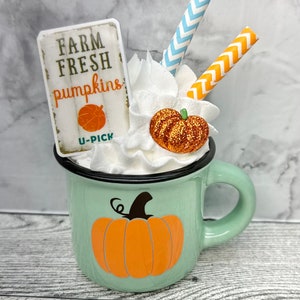 May include: A mint green enamel mug with a black rim and handle, decorated with an orange pumpkin graphic. The mug is filled with whipped cream, a glittery pumpkin, and two straws. A sign that reads "FARM FRESH pumpkins" is also included.