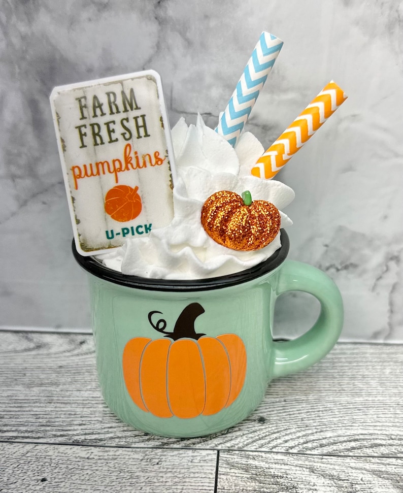 May include: A mint green enamel mug with a black rim and handle, decorated with an orange pumpkin graphic. The mug is filled with whipped cream, a glittery pumpkin, and two straws. A sign that reads "FARM FRESH pumpkins" is also included.