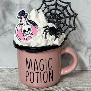 May include: A pink ceramic mug with a black rim and handle. The mug has a black and white graphic that says "Magic Potion". The mug is decorated with a white whipped cream topping, a black spider, and a pink potion bottle with a skull and crossbones graphic. The potion bottle is topped with a black spider web.