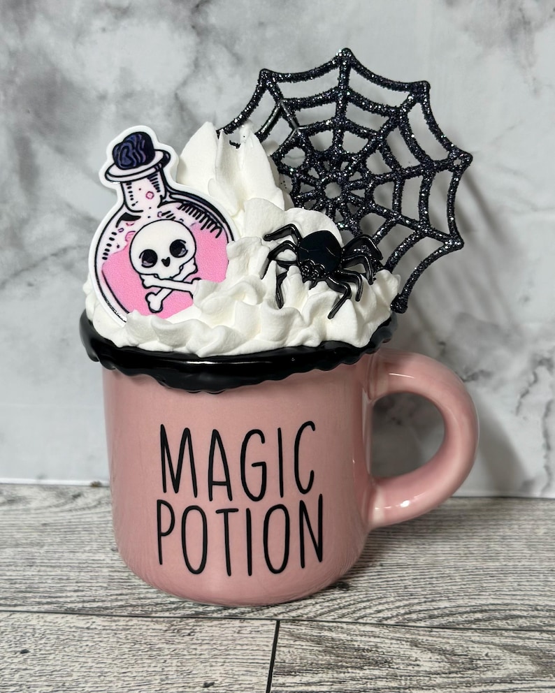May include: A pink ceramic mug with a black rim and handle. The mug has a black and white graphic that says "Magic Potion". The mug is decorated with a white whipped cream topping, a black spider, and a pink potion bottle with a skull and crossbones graphic. The potion bottle is topped with a black spider web.