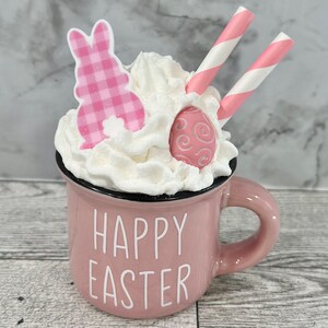 Op de afbeelding: Een roze keramische mok met witte slagroom, een roze konijn en een roze en wit gestreepte rietje. De mok heeft de tekst "Happy Easter" aan de voorkant.