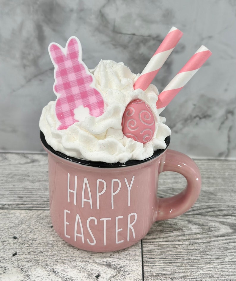 Op de afbeelding: Een roze keramische mok met witte slagroom, een roze konijn en een roze en wit gestreepte rietje. De mok heeft de tekst "Happy Easter" aan de voorkant.