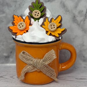 May include: An orange ceramic mug with a jute bow tied around it. The mug is topped with white whipped cream and decorated with felt leaves in orange, yellow, and green, each adorned with a wooden button. A fall-themed decorative item.
