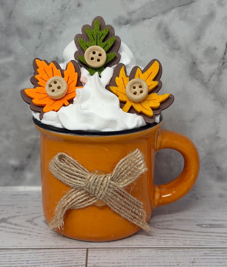 May include: An orange ceramic mug with a jute bow tied around it. The mug is topped with white whipped cream and decorated with felt leaves in orange, yellow, and green, each adorned with a wooden button. A fall-themed decorative item.