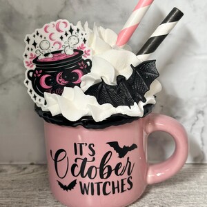 May include: Pink ceramic mug with a black rim and handle. The mug has a white whipped cream topping with a black bat and pink and white striped straws. The mug has a black graphic that says "It's October Witches" with a black bat on either side of the text. There is a pink and black sticker on the mug with a cauldron and the word "Hello".