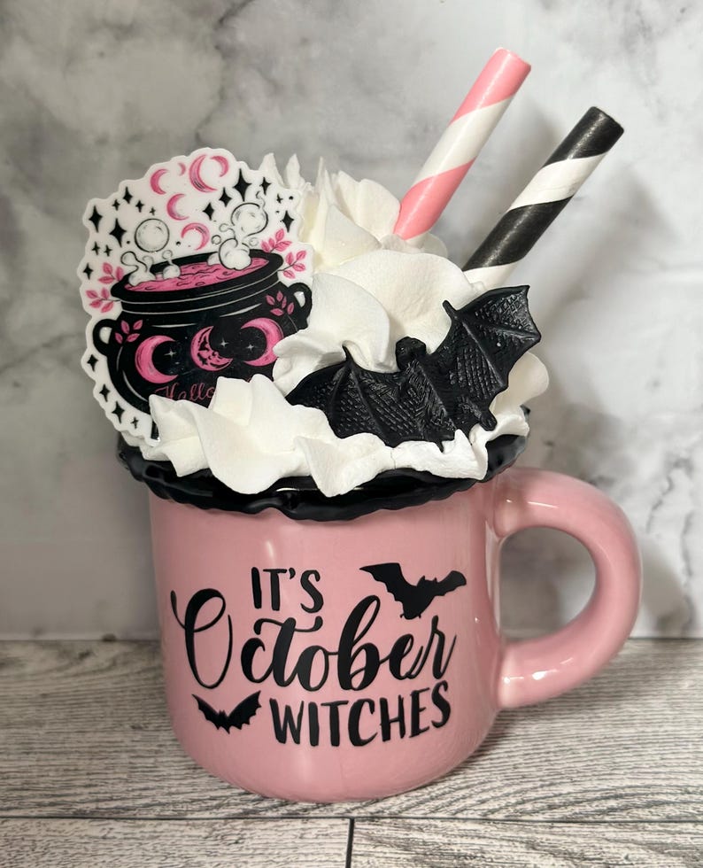 May include: Pink ceramic mug with a black rim and handle. The mug has a white whipped cream topping with a black bat and pink and white striped straws. The mug has a black graphic that says "It's October Witches" with a black bat on either side of the text. There is a pink and black sticker on the mug with a cauldron and the word "Hello".